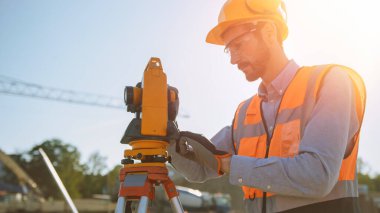 İnşaat İşçisi İnşaat Alanındaki Yatay ve Dikey Düzlemlerdeki Açıları Ölçmek İçin Optik Araştırma Aracı Kullanıyor. Bina için Sert Şapka Yapımında İşçi Projeksiyonu.