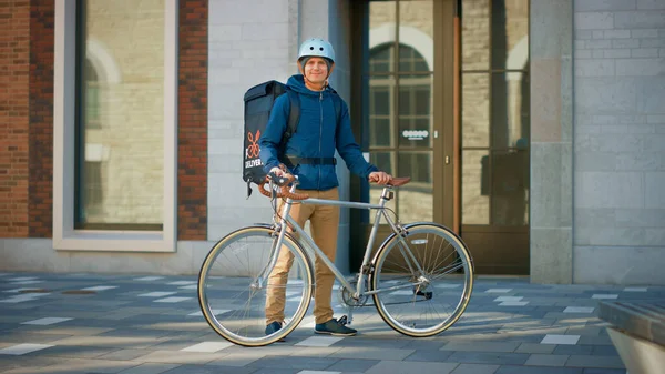 在时尚的现代城区，英俊的送货员穿着热背包和安全头盔站在他的自行车旁边。《微笑信使安全送餐令》的肖像 — 图库照片