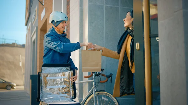 Happy Food Delivery Man Wearing Thermal Backpack on a Bike Delivers Restaurant Order to a Female Customer. Courier Delivers Takeaway Lunch to Gorgeous Girl in Modern City District Office Building