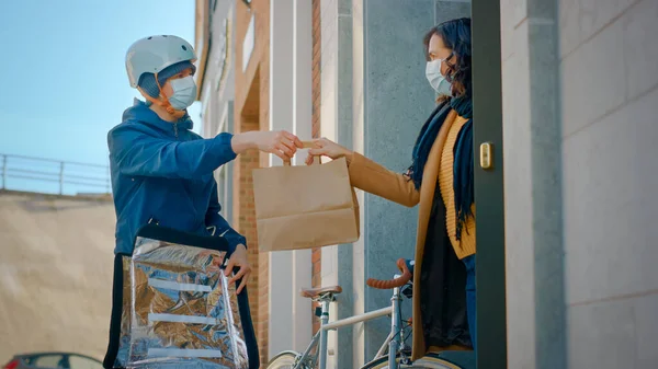 Food Delivery Man φορώντας προστατευτική μάσκα προσώπου και θερμική σακίδιο σε ένα ποδήλατο παραδίδει την παραγγελία σε μια μασκοφόρο θηλυκό πελάτη. Ταχυμεταφορέας παραδίδει φαγητό. Καραντίνα, κοινωνική απόσταση — Φωτογραφία Αρχείου