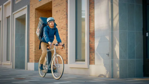 英俊快乐的送餐人穿着热背包骑着自行车穿过时尚的现代城区。《微笑信使安全送餐令》开枪射击. — 图库照片