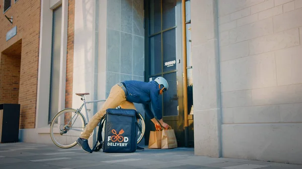 食物派递员穿着保暖背包骑单车送递餐厅订单，把它放在门下。概念：无接触、无接触、无社会疏离、无自我隔离 — 图库照片