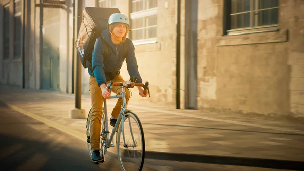 快乐食品快递员穿着保暖背包骑着自行车为客户和顾客送货上门。现代城市阳光灿烂,城市建筑时尚时尚.用温滤器射击. — 图库照片