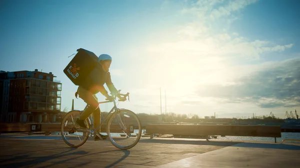 快乐食品快递员穿着保暖背包骑着自行车为顾客运送订单和包装。近岸城市阳光明媚，建筑别致. — 图库照片