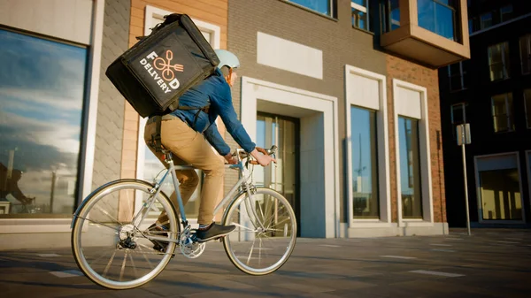 Happy Food Delivery Courier Φορώντας Θερμική Σακίδιο Οδηγεί Ένα Ποδήλατο Στο Δρόμο Για Να Παραδώσει Παραγγελίες και Πακέτα Για Πελάτες. Ηλιόλουστη μέρα στην πόλη με κομψά αστικά κτίρια. — Φωτογραφία Αρχείου