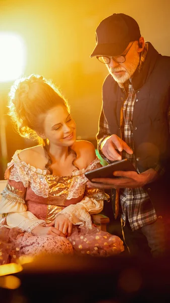 On Period Costume Drama Film Set: Beautiful Smiling Actress Wearing Renaissance Dress Sitting on a Chair Listens to Movie Director Explaining to Her Scene Context. High Budget Period Drama.