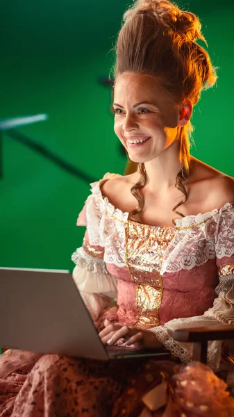 On Period Costume Drama Film Set: Beautiful Smiling Actress Wearing Renaissance Dress, Sitting on a Chair Using Laptop Computer with Green Screen in the Background.