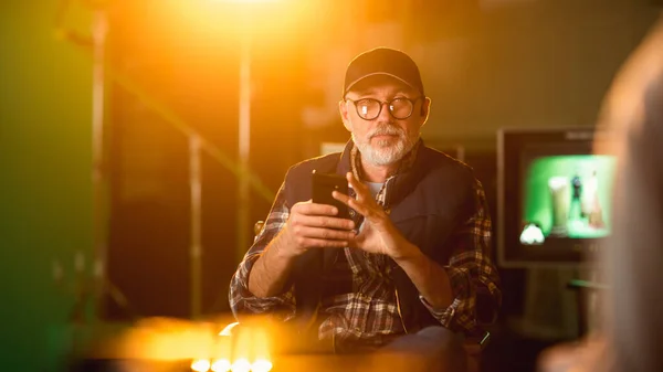 Réalisateur éminent assis sur une chaise pendant une pause à l'aide d'un téléphone intelligent. Sur le plateau de tournage studio avec équipement haut de gamme tournage de l'équipage professionnel film à budget élevé. — Photo