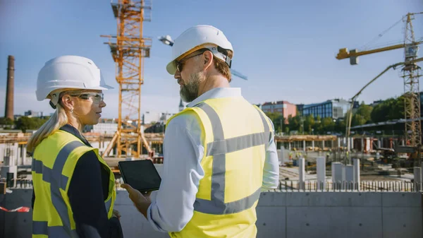 Ingeniero civil masculino y joven arquitecta de edificios femeninos Utilice un Tablet Computer en un sitio de construcción de la ciudad. Hablan sobre el futuro del desarrollo y la planificación de bienes raíces. Usar sombreros duros de seguridad. — Foto de Stock