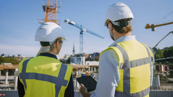 Ingeniero civil masculino y joven arquitecta de edificios femeninos Utilice un Tablet Computer en un sitio de construcción de la ciudad. Hablan sobre el futuro del desarrollo y la planificación de bienes raíces. Usar sombreros duros de seguridad. — Foto de Stock