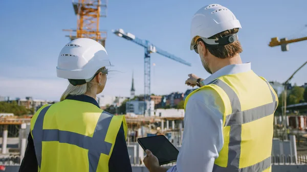 Ingeniero civil masculino y joven arquitecta de edificios femeninos Utilice un Tablet Computer en un sitio de construcción de la ciudad. Hablan sobre el futuro del desarrollo y la planificación de bienes raíces. Usar sombreros duros de seguridad. —  Fotos de Stock