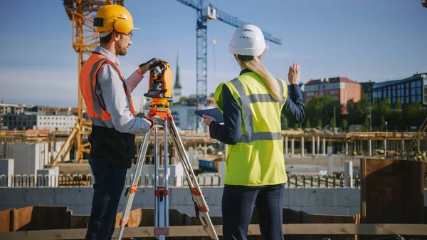 Operaio edile che utilizza lo strumento ottico di rilevamento della teodolite per misurare gli angoli in piani orizzontali e verticali sul cantiere. Ingegnere e architetto utilizzando tavoletta accanto al geometra. — Foto Stock