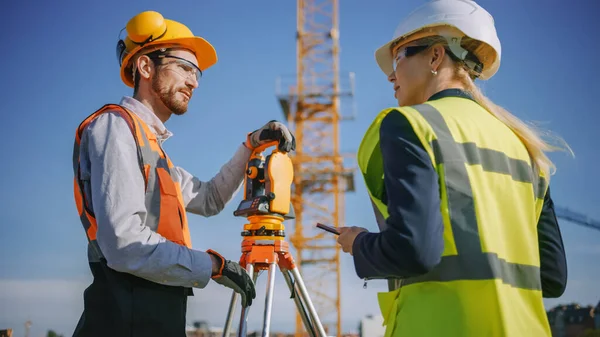 Operaio edile che utilizza lo strumento ottico di rilevamento della teodolite per misurare gli angoli in piani orizzontali e verticali sul cantiere. Ingegnere e architetto utilizzando tavoletta accanto al geometra. — Foto Stock