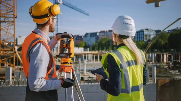 Operaio edile che utilizza lo strumento ottico di rilevamento della teodolite per misurare gli angoli in piani orizzontali e verticali sul cantiere. Ingegnere e architetto utilizzando tavoletta accanto al geometra. — Foto Stock