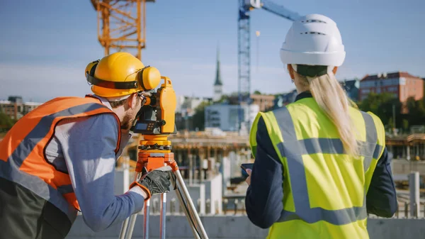 Pracownik budowlany wykorzystujący teodolitowy przyrząd pomiarowy do pomiaru kątów w płaszczyznach poziomych i pionowych na placu budowy. Inżynier i architekt korzystający z tabletu obok geodety. — Zdjęcie stockowe