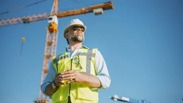El Confident Bearded Head Civil Engineer-Architect in Sunglasses está parado afuera en un sitio de construcción en un día soleado y brillante. El hombre está usando un sombrero duro, camisa, pantalones vaqueros y un chaleco de seguridad amarillo. — Foto de Stock