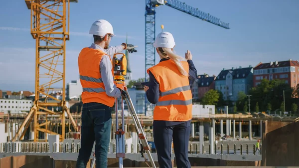 Operaio edile che utilizza lo strumento ottico di rilevamento della teodolite per misurare gli angoli in piani orizzontali e verticali sul cantiere. Ingegnere e architetto utilizzando tavoletta accanto al geometra. — Foto Stock