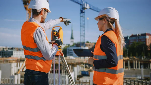 Operaio edile che utilizza lo strumento ottico di rilevamento della teodolite per misurare gli angoli in piani orizzontali e verticali sul cantiere. Ingegnere e architetto utilizzando tavoletta accanto al geometra. — Foto Stock