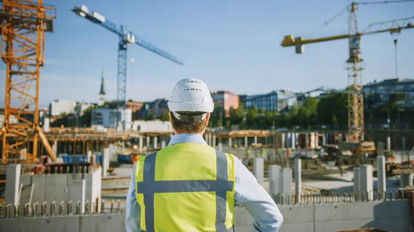 El Confident Bearded Head Civil Engineer-Architect in Sunglasses está parado afuera con su espalda a la cámara en un sitio de construcción en un día brillante. El hombre lleva un sombrero duro, una camisa y un chaleco de seguridad. — Foto de Stock