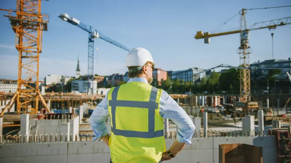 El Confident Bearded Head Civil Engineer-Architect in Sunglasses está parado afuera con su espalda a la cámara en un sitio de construcción en un día brillante. El hombre lleva un sombrero duro, una camisa y un chaleco de seguridad. — Foto de Stock