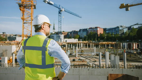 El Confident Bearded Head Civil Engineer-Architect in Sunglasses está parado afuera con su espalda a la cámara en un sitio de construcción en un día brillante. El hombre lleva un sombrero duro, una camisa y un chaleco de seguridad. — Foto de Stock