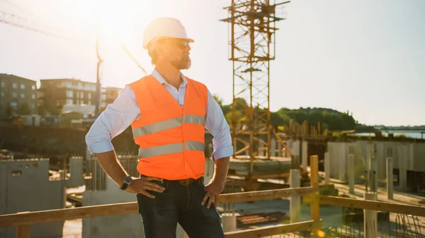 Confiado Jefe de Ingeniero Civil-Arquitecto Barbudo en Gafas de Sol se encuentra en un sitio de construcción en un día soleado y brillante. El hombre está usando un sombrero duro, camisa, pantalones vaqueros y un chaleco de seguridad naranja. — Foto de Stock
