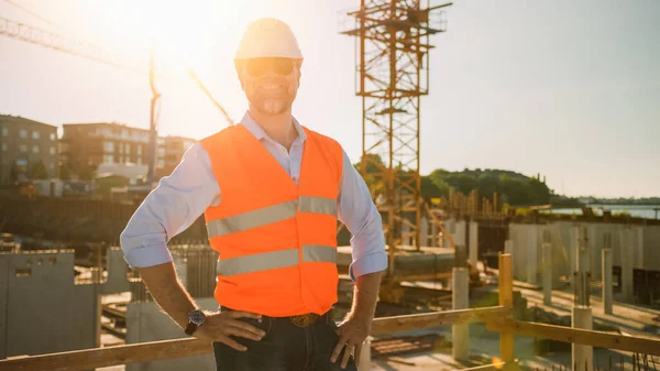 El Confident Bearded Head Civil Engineer-Architect in Sunglasses está sonriendo en la cámara en un sitio de construcción en un día soleado y brillante. El hombre está usando un sombrero duro, camisa, pantalones vaqueros y un chaleco de seguridad naranja. — Foto de Stock
