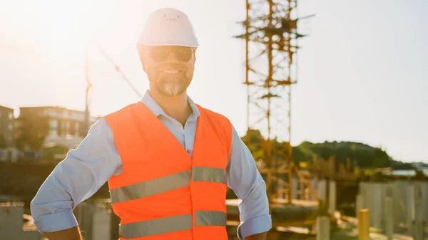 Confident Bearded Head Civil Engineer-Architect in Sunglasses is Smiling on Camera in a Construction Site on a Sunny Bright Day. Man is Wearing a Hard Hat, Shirt, Jeans and an Orange Safety Vest.