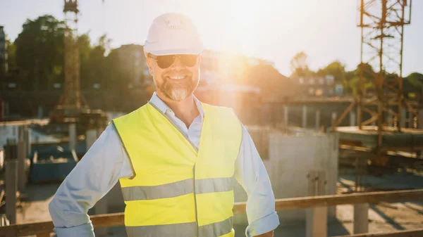 Σίγουρος Γενειοφόρος Επικεφαλής Πολιτικός Μηχανικός-Αρχιτέκτονας στην Sunglasses χαμογελά στην κάμερα σε ένα εργοτάξιο σε μια ηλιόλουστη φωτεινή ημέρα. Ο άνθρωπος φοράει ένα σκληρό καπέλο, πουκάμισο, τζιν και ένα κίτρινο γιλέκο ασφαλείας. — Φωτογραφία Αρχείου