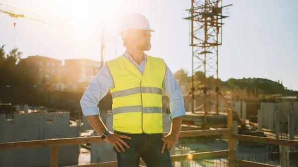 Confiado Jefe de Ingeniero Civil-Arquitecto Barbudo en Gafas de Sol se encuentra en un sitio de construcción en un día soleado y brillante. El hombre está usando un sombrero duro, camisa, pantalones vaqueros y un chaleco de seguridad amarillo. — Foto de Stock