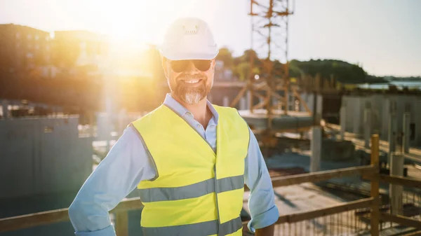 El Confident Bearded Head Civil Engineer-Architect in Sunglasses está sonriendo en la cámara en un sitio de construcción en un día soleado y brillante. El hombre está usando un sombrero duro, camisa, pantalones vaqueros y un chaleco de seguridad amarillo. — Foto de Stock