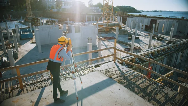 Trabajador de la construcción utilizando el instrumento óptico de levantamiento de teodolitos para medir ángulos en planos horizontales y verticales en el sitio de construcción. Trabajador en casco duro haciendo proyecciones para el edificio. — Foto de Stock