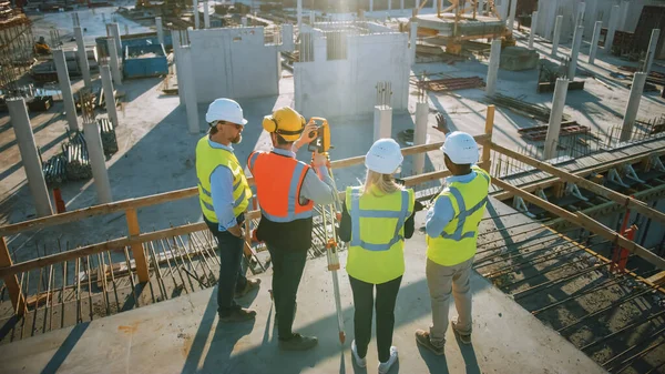 Operaio edile che utilizza lo strumento ottico di rilevamento della teodolite per misurare gli angoli in piani orizzontali e verticali sul cantiere. Ingegneri e architetti discutono i piani accanto al geometra. — Foto Stock