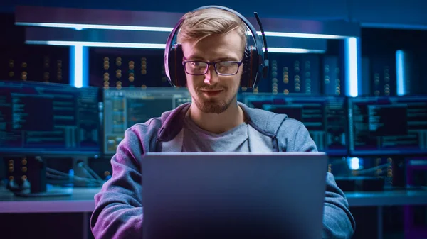 软件开发人员Hacker Gamer戴着眼镜和耳机坐在办公桌前，在笔记本电脑上玩的肖像。背景下的多显示黑暗高科技环境. — 图库照片