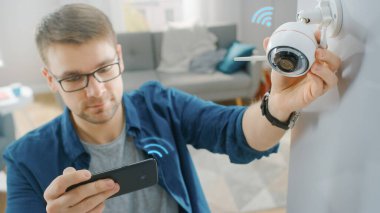Gözlüklü Genç Adam Mavi Gömlek Giyiyor. Evdeki Beyaz Duvarda İki Antenli Modern Wi-Fi Gözetleme Kamerasını ayarlıyor. Akıllı telefonundaki video yayınını kontrol ediyor..