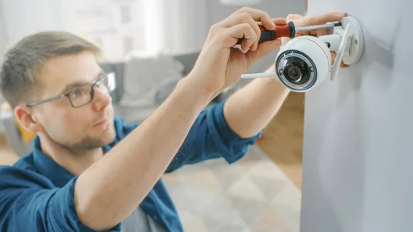 Jovem de óculos vestindo uma camisa azul está fodendo uma câmera de vigilância Wi-Fi moderna com duas antenas em uma parede branca em casa. — Fotografia de Stock