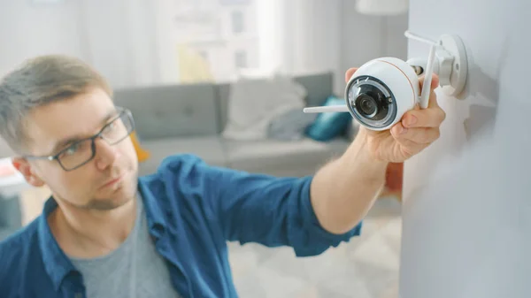 Jovem de óculos vestindo uma camisa azul está fodendo uma câmera de vigilância Wi-Fi moderna com duas antenas em uma parede branca em casa. — Fotografia de Stock