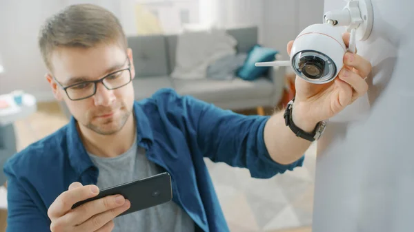 O jovem de óculos vestindo uma camisa azul está ajustando uma câmera de vigilância Wi-Fi moderna com duas antenas em uma parede branca em casa. Ele está verificando o feed de vídeo em seu Smartphone. — Fotografia de Stock