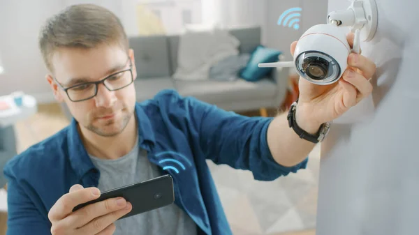 Nuori mies laseissa yllään sininen paita säätää modernia Wi-Fi-valvontakameraa, jossa on kaksi antennia valkoisella seinällä kotona. Hän tarkistaa videosyötteen älypuhelimellaan.. — kuvapankkivalokuva