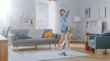 Kot Gömlekli ve Şortlu Genç Güzel Kadın Kafasında Müzik Dinliyor, Evde Halıyı Temizlerken Dans Ediyor ve Vakum Temizliyor. Kablosuz Süpürge kullanıyor. O mutlu ve neşeli..