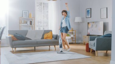 Kot gömlekli, şortlu dans eden ve evdeki rahat bir odada halı temizleyen genç ve güzel bir kadının fotoğrafı. Modern Kablosuz Süpürge kullanıyor. O mutlu..