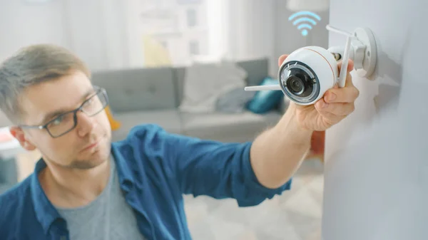 Junger Mann mit Brille und blauem Hemd justiert zu Hause eine moderne WLAN-Überwachungskamera mit zwei Antennen an einer weißen Wand. — Stockfoto