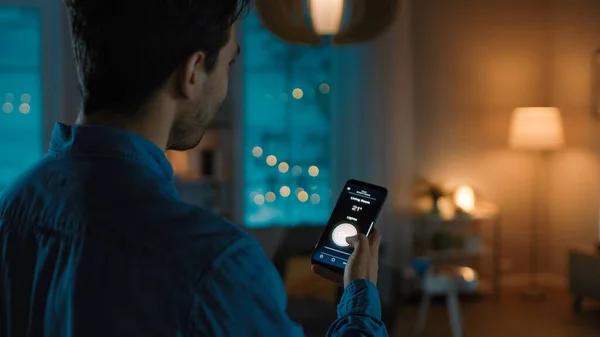 Joven hombre guapo está sosteniendo Smartphone con la aplicación activa Smart Home. Persona está tocando la pantalla para encender las luces en la habitación. Su Acogedora Noche en el Apartamento. — Foto de Stock