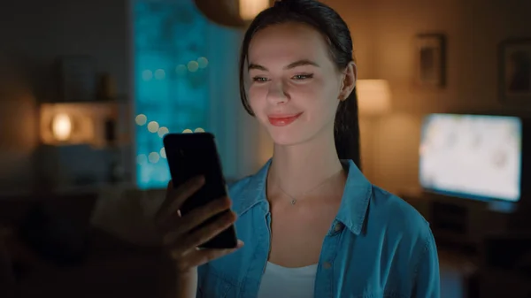 Porträt einer jungen schönen Frau, die Smartphone benutzt, im Internet surft, soziale Netzwerke checkt und Smart-Home-Anwendungen nutzt, während sie zu Hause steht. Gemütlicher Abend. — Stockfoto