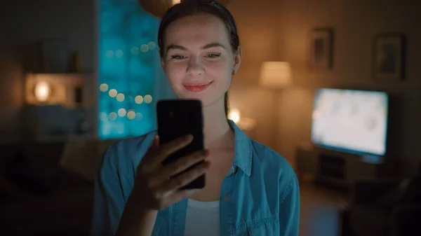 Porträt einer jungen schönen Frau, die Smartphone benutzt, im Internet surft, soziale Netzwerke checkt und Smart-Home-Anwendungen nutzt, während sie zu Hause steht. Gemütlicher Abend. — Stockfoto