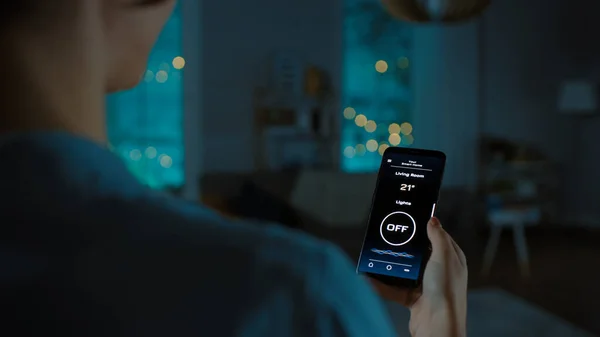 Fermer Vue d'ensemble d'un Smartphone avec application Active Smart Home. Jeune femme tapote sur l'écran pour allumer la lumière dans la pièce. Sa soirée confortable dans l'appartement. — Photo