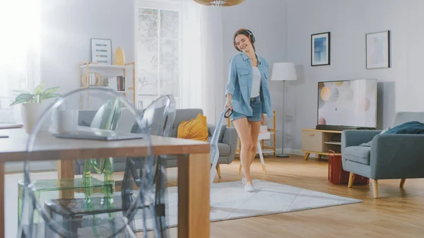 Young Beautiful Woman in Jeans Shirt and Shorts écoute de la musique sur ses headpones, danse et vide un tapis dans une chambre confortable à la maison. Elle utilise un aspirateur sans fil. Elle est heureuse et joyeuse. — Photo