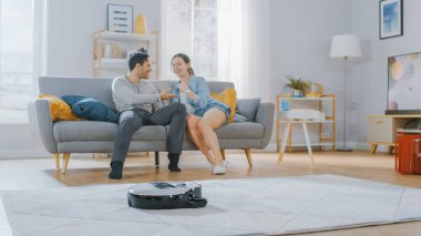 Akıllı Robot Elektrikli Süpürge Halıdan Toz Emici. Güzel bir çift bir koltukta oturuyor ve arka planda konuşuyor. Teknolojik Ev Aletleri Cihazı Onları Geçirir.