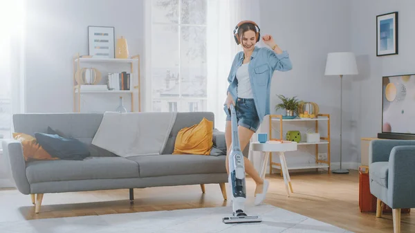 Shot of a Young Beautiful Woman in Jeans Shirt and Shorts Dancing and Vacuum Cleaning a Carpet in a Cozy Room at Home (en inglés). Utiliza un vacío inalámbrico moderno. Ella es feliz.. — Foto de Stock
