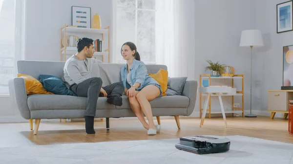 Aspirateur robot intelligent aspirant la poussière d'un tapis. Beau couple est assis sur un canapé et parle en arrière-plan. Appareil électroménager technologique se déplace les passé. — Photo